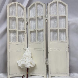 May include: A three-panel folding room divider in distressed off-white wood with arched tops and six window panes per panel, featuring white sheer curtains behind the panes. A small white lace parasol with a dark red handle rests against the center panel.