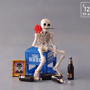 May include: A 1/12 scale articulated skeleton figure sitting on a blue plastic beer crate, holding a red rose in its mouth. The scene includes three brown glass beer bottles and a small gold-framed picture of a person with a green apple face.