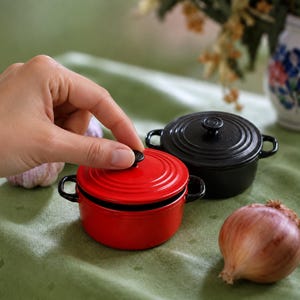 May include: Two small, round, enameled cast iron Dutch ovens with lids and side handles. One pot is bright red and the other is black. Both feature a circular ridged design on the lid and a small central knob for lifting the lid off the pot.