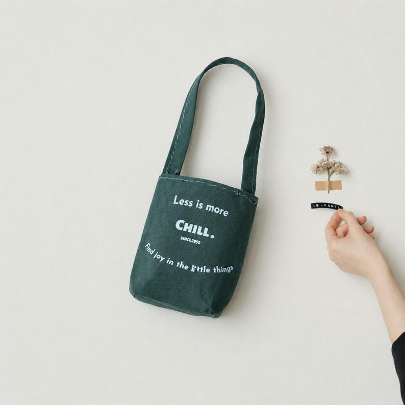Green miniature canvas tote bag displayed on a wall with small flower decor and a hand for scale.