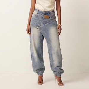 May include: Light blue denim jeans featuring a high-waisted, asymmetrical wrap-front design with a layered waistband, a tan leather-like patch on the front, and a relaxed, loose-fitting silhouette that tapers toward the ankles with cuffed hems.