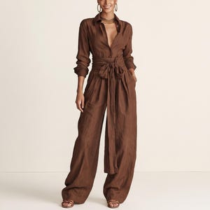 Op de afbeelding: Een donkerbruine jumpsuit met lange mouwen, voorzien van een overhemdtop met kraag en knopen, een brede geïntegreerde ceintuur en een wijde broek. Het kledingstuk is gemaakt van een gestructureerde stof en bevat bijpassende bruine slippers.