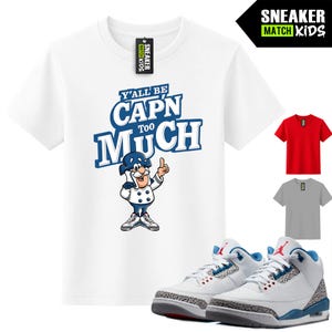 May include: A white short-sleeve t-shirt featuring the blue text Y'ALL BE CAP'N TOO MUCH above a cartoon character wearing a blue hat and white uniform. The shirt includes a black neck tag and is displayed alongside a pair of white, blue, and grey sneakers.