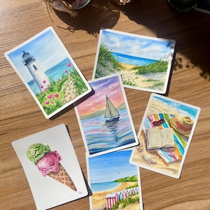 Peut inclure: Une collection de six peintures rectangulaires à l'aquarelle représentant des scènes côtières, dont un phare, un sentier de plage, un voilier au coucher du soleil, une serviette de plage avec un livre et un chapeau, des cabines et une glace.