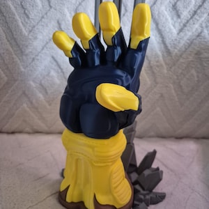 May include: A 3D printed sculpture of a stylised hand wearing a dark blue and yellow glove. The hand is positioned with fingers extended upwards, featuring a yellow cuff at the wrist and a base decorated with dark grey jagged rock-like fragments.
