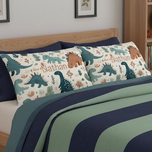 May include: Two rectangular pillowcases featuring a cream background with a repeating pattern of various dinosaurs in shades of teal, dark green, and brown, interspersed with foliage. The name Nathan is printed in a brown script font on each.