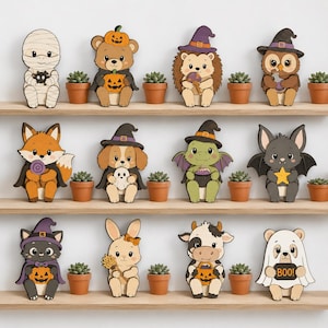 Puede incluir: Una colección de doce figuras de animales de madera vestidas con disfraces de Halloween, incluyendo una momia, un oso con calabazas, un erizo, un búho, un zorro, un perro, un dragón, un murciélago, un gato, un conejo, una vaca y un fantasma con un cartel BOO.