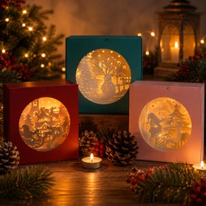 May include: Three square boxes in red, dark green, and light pink, each featuring a circular cutout window. Inside each window is a backlit paper-cut scene depicting winter landscapes with silhouettes of a snowman, Santa in a sleigh, and a deer.