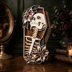 May include: A multi-layered wooden wall art piece shaped like a coffin, featuring a central skeleton torso and skull. The design is accented with layered floral motifs in shades of deep red, dark green, and natural wood tones, with small star details.