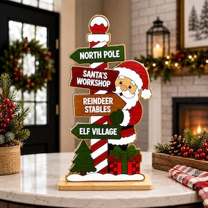 May include: A decorative wooden sign featuring a red and white striped pole with four directional arrows labelled North Pole, Santa's Workshop, Reindeer Stables, and Elf Village. A cartoon Santa Claus stands beside the pole next to a gift box.