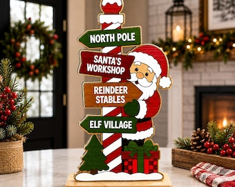 May include: A wooden tabletop sign featuring a red and white striped pole with four directional signs reading North Pole, Santa's Workshop, Reindeer Stables, and Elf Village. A Santa Claus figure stands beside the pole above a gift box.