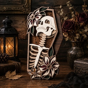May include: A multi-layered wooden wall art piece shaped like a coffin, featuring a light-colored skeleton torso and skull surrounded by dark-toned floral accents. The design includes intricate cutouts of flowers and leaves with deep red and green details.