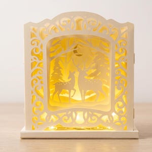 May include: A cream-colored, layered paper light box featuring a laser-cut frame with intricate scrollwork. The interior displays a silhouette scene of two deer standing in a forest with pine trees and a crescent moon, illuminated by a warm yellow light.