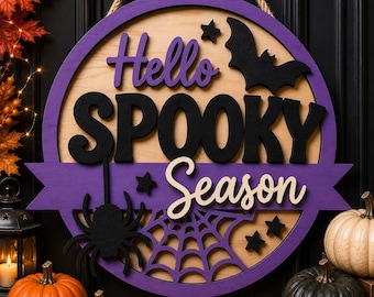May include: A round wooden wall sign featuring the text Hello SPOOKY Season in purple, black, and cream. The design includes a black bat, a black spider, a spider web cutout, and small black stars. A thick jute rope is attached for hanging.