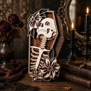 May include: A multi-layered wooden wall art piece shaped like a coffin, featuring a light-colored skull and rib cage skeleton design. The artwork is decorated with dark red and green floral accents and small star engravings on a dark wood background.