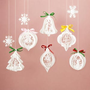 Puede incluir: Una colección de adornos de papel blanco colgados de hilos, incluyendo copos de nieve, una campana, un árbol y formas esféricas. Algunos presentan recortes de un muñeco de nieve, un ciervo y el texto Merry Christmas, con lazos verdes, rosas, rojos y amarillos.