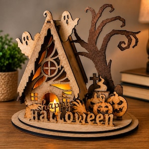 May include: A laser-cut wooden Halloween decoration featuring a house with a glowing light inside, two ghosts, a bare tree, tombstones, and two jack-o'-lanterns. The base includes the text "happy HALLOWEEN" in a cutout script and block font.