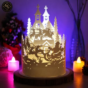 May include: A cylindrical white paper lantern featuring intricate laser-cut silhouettes of Santa Claus in a sleigh pulled by reindeer, flying over a village with a church and pine trees. The lantern is illuminated from within, casting a warm glow.