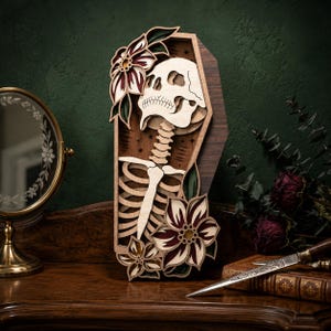 May include: A multi-layered wooden wall art piece shaped like a coffin, featuring a light-colored skull and ribcage skeleton design. The artwork is decorated with dark red and green floral accents and small star motifs on a dark wood background.