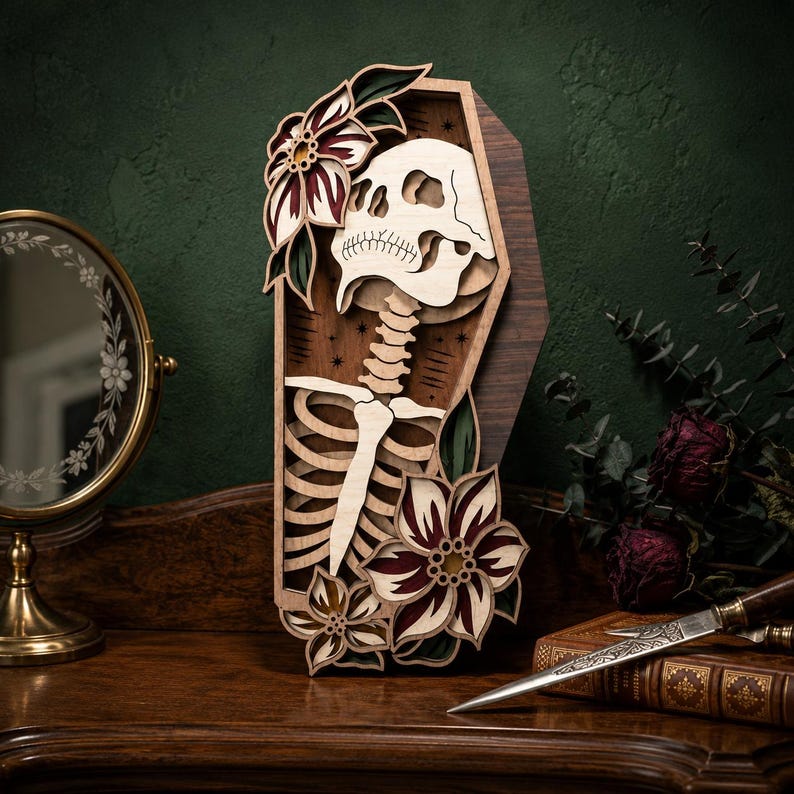 May include: A multi-layered wooden wall art piece shaped like a coffin, featuring a light-colored skull and ribcage skeleton design. The artwork is decorated with dark red and green floral accents and small star motifs on a dark wood background.