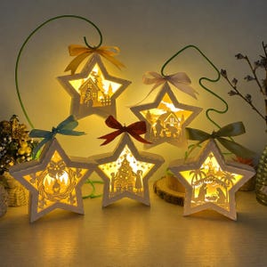 May include: Five star-shaped wooden lanterns with warm internal lighting, each featuring intricate cut-out holiday scenes including a snowman, a nativity, and a decorated tree. Each lantern is topped with a colorful ribbon and a metal hook.