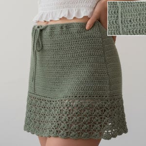 May include: A sage green crochet skirt featuring a drawstring waist and a solid stitch pattern on the upper section, transitioning into an intricate, openwork floral lace border at the hem. The inset shows a close-up of the textured yarn stitches.