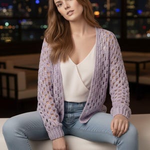 May include: A light purple open-front cardigan featuring a loose crochet stitch pattern with long sleeves and ribbed cuffs, worn over a white sleeveless top and light blue denim jeans. The garment has a relaxed fit and a textured, airy appearance.