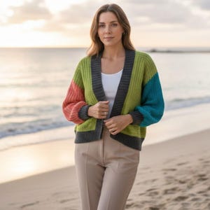 May include: A color-blocked open-front cardigan featuring a lime green body, a coral sleeve, a teal sleeve, and dark gray ribbed trim along the neckline, cuffs, and hem. The knit garment is worn over a plain white top and beige trousers.