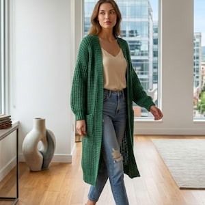 Può includere: Un lungo cardigan verde smeraldo aperto sul davanti, caratterizzato da una trama a maglia grossa a nido d'ape, maniche lunghe e due grandi tasche applicate. Il capo è indossato sopra una canotta beige e jeans azzurri consumati con uno strappo.