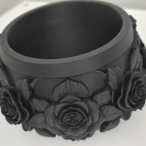 May include: A matte black cylindrical container featuring a raised, three-dimensional relief pattern of blooming roses and leaves encircling the entire exterior surface. The object has a smooth, thick rim and a deep, hollow interior cavity.