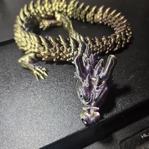 May include: A 3D printed articulated dragon figurine featuring a color-shifting finish that transitions between metallic gold and deep purple. The creature has a detailed head with horns and a long, segmented body with sharp, overlapping scales.