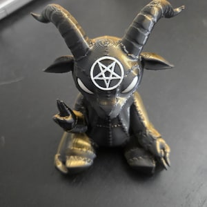 May include: A 3D-printed figurine of a seated goat-like creature with large curved horns, pointed ears, and white eyes. The figure is dark gray with a visible layered texture and features a white pentagram symbol centered on its forehead.