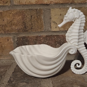 May include: A matte white ceramic dish shaped like a scalloped seashell, featuring a detailed seahorse figure attached to one side. The seahorse is upright with a curled tail, and the bowl has a smooth, curved interior with deep, flowing ridges.