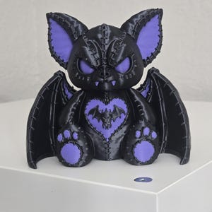 May include: A black and purple bat figurine featuring stitched detailing, large pointed ears, and a heart-shaped emblem on the chest with a smaller bat graphic. The creature has wide, glowing purple eyes and small fangs, with wings extended to the sides.