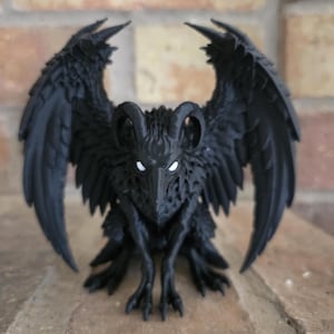 Puede incluir: Una figura de color negro mate de una criatura alada con cuernos curvados y garras afiladas. La criatura presenta texturas de plumas detalladas en sus alas y cuerpo, con ojos blancos brillantes que contrastan con el material oscuro y sólido.