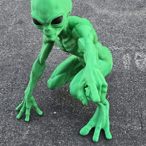 May include: A bright green alien figurine in a crouching position with an elongated head, large black oval eyes, and long slender limbs. The figure has detailed muscular definition and four-fingered hands reaching forward.