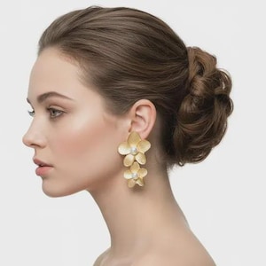 May include: A pair of gold-toned drop earrings featuring two stacked flower-shaped components. Each flower has five textured petals and a small, round, white pearl-like bead centered in the middle of the bloom, creating a layered floral design.