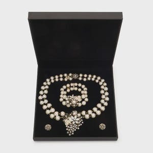May include: A jewellery set consisting of a double-strand pearl necklace, a double-strand pearl bracelet, and two matching stud earrings. The pieces feature white pearls and floral-shaped metal accents with clear crystals, presented in a black box.