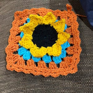May include: A square crochet granny square featuring a central sunflower motif with a black center and yellow petals. The sunflower is layered over a bright blue crochet base, all framed by a textured border of burnt orange yarn stitches.