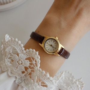 Könnte beinhalten: Eine goldfarbene Armbanduhr mit braunem Lederarmband wird am Handgelenk getragen. Die Uhr hat ein weißes Zifferblatt mit schwarzen Ziffern und Zeigern. Das Handgelenk befindet sich neben einem weißen Spitzensaum.