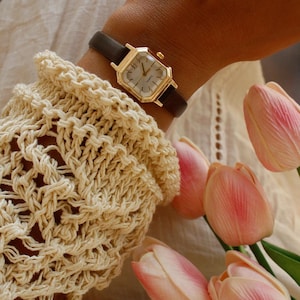May include: A gold-toned rectangular watch with a brown leather strap is displayed. The watch has a light-colored face with black Roman numerals. The watch is worn on a wrist, next to a cream-colored crocheted sleeve and pink tulips.