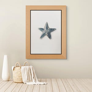 May include: A framed wall art piece featuring a blue starfish centered on a white background, enclosed in a light-toned wooden frame. The artwork is displayed above a woven basket containing a white cloth and a tall, smooth white ceramic vase.