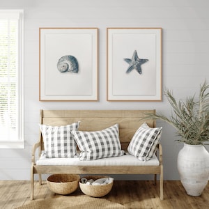 May include: Two framed wall art prints featuring a blue seashell and a blue starfish on white backgrounds, displayed above a wooden bench with three gray and white checkered pillows, two woven baskets, and a large white ceramic vase.