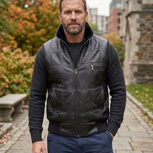 Op de afbeelding: Een zwart leren vest met een gewatteerd patroon, een zwarte bontkraag en een ritssluiting. Het wordt gedragen over een donkergrijze trui en gecombineerd met een blauwe broek. De achtergrond toont een park met bomen en een stenen gebouw.