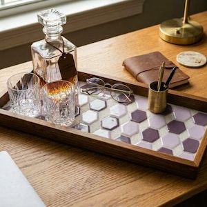 May include: A rectangular wooden serving tray featuring a mosaic of purple and white hexagonal tiles. The tray holds a clear glass decanter with a stopper, two matching crystal-style drinking glasses, a pair of round eyeglasses, and a brass pen holder.