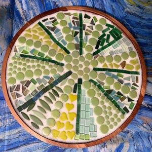 May include: A circular wooden tray featuring a mosaic design made of various green, yellow, and light blue glass pieces. The mosaic includes round, triangular, and heart-shaped tiles arranged in a radial pattern on a white base within the frame.