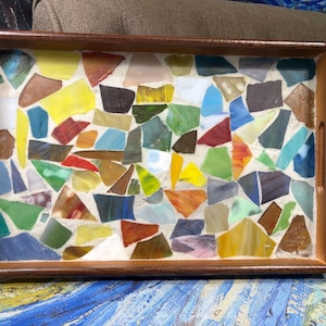 May include: A rectangular wooden serving tray featuring a mosaic inlay made of multicolored stained glass pieces. The glass fragments in shades of yellow, blue, green, red, and brown are set in a white grout, framed by a dark wood border with handles.