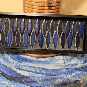 May include: A rectangular black wooden frame containing a series of alternating blue and black ceramic leaf-shaped tiles arranged in a repeating pattern against a dark textured background, creating a structured and geometric wall art piece.