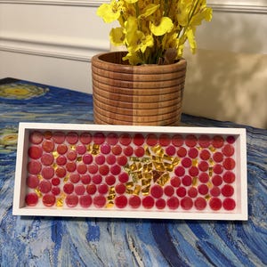 May include: A rectangular white tray filled with a mosaic of small, circular, translucent red glass pieces and scattered, irregular gold-colored metallic shards embedded in a clear resin base, creating a vibrant, textured, and reflective surface.