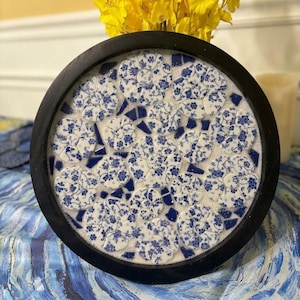 May include: A circular wall art piece featuring a black frame surrounding a mosaic design. The mosaic is composed of white and blue floral patterned ceramic shards arranged in circular flower shapes, interspersed with small solid dark blue pieces.